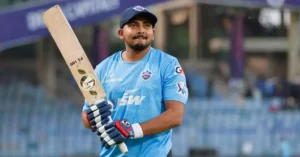 The image depicts a cricket player holding a cricket bat, wearing a blue jersey with "JSW" written on it, and representing a team in a cricket stadium. The player looks determined and is wearing gloves and a cap.