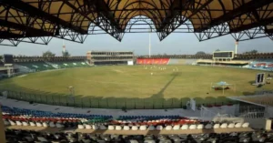 Wide-angle view of Gaddafi Stadium in Lahore, showcasing its iconic roof structure, empty colorful seating, and cricket field under preparation, with a few players practicing in the distance.